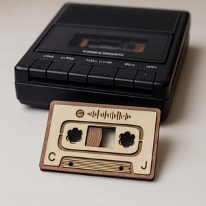 Peut inclure: Un lecteur de cassettes noir avec un design de cassette en bois. La cassette en bois a une face marron clair avec une bordure marron foncé et présente un graphique d'onde sonore et les lettres "C" et "J".