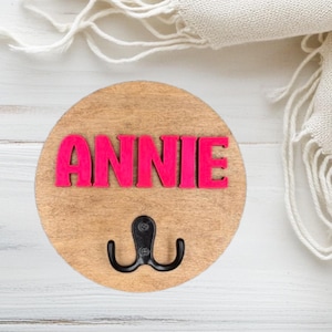 May include: A circular wooden wall hook featuring the name "ANNIE" in vibrant pink lettering. A black double hook is attached below the name. The wood has a light brown finish.