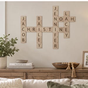May include: A wall-mounted display of light wood square tiles arranged like a crossword puzzle, featuring the names Jacob, Bailey, Christine, Jennifer, Noah, and Lacey in black letters with small numerical values printed in the bottom right corners.