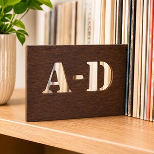 以下が含まれることがあります： 「A-D」の文字が切り抜かれた濃い茶色の木製サイン。明るい表面が見えます。木製の棚に置かれ、白い鉢植えの植物と背景にレコードの山があります。