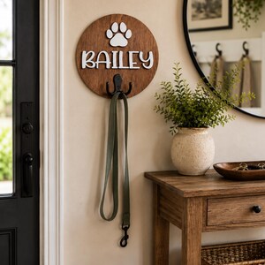Könnte beinhalten: Ein rundes Wandschild aus Holz mit einem weißen Pfotenabdruck-Symbol und dem Namen BAILEY in weißen, erhabenen Buchstaben. Ein schwarzer Metallhaken ist unten in der Mitte des Schildes befestigt und hält eine lange olivgrüne Tierleine.