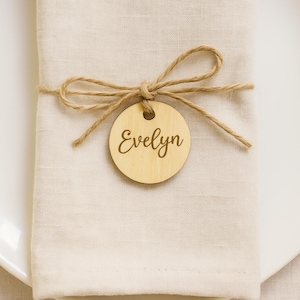May include: A light beige linen napkin is tied with twine, featuring a wooden round tag. The tag is engraved with the name "Evelyn" in a cursive font. The napkin is placed on a white plate, creating a neutral and elegant aesthetic.