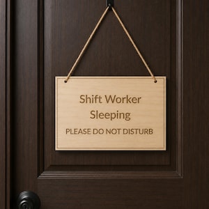 May include: A rectangular light-colored wooden sign hanging from a brown twine cord. The sign features the engraved text Shift Worker Sleeping and PLEASE DO NOT DISTURB in a dark brown font, with a thin border line around the edge.