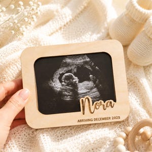 Op de afbeelding: Een houten frame met een echografiebeeld. Het frame heeft de naam "Nora" en de tekst "ARRIVING DECEMBER 2025" gegraveerd. De afbeelding wordt omringd door babyartikelen, waaronder witte babyslofjes en een houten speelgoed.