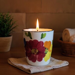 Peut inclure: Une bougie allumée dans un pot en céramique blanc orné de fleurs colorées. Les fleurs sont rouges, jaunes et oranges. La bougie repose sur un tissu beige. Une petite plante et des serviettes roulées sont en arrière-plan.