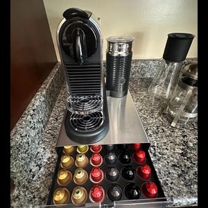 Può includere: Una macchina per caffè espresso nera e argento con un montalatte e un cassetto pieno di capsule di caffè colorate. La macchina è su un ripiano in acciaio inossidabile, accanto a un contenitore di vetro con coperchio nero.