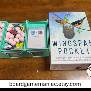 May include: Wingspan Pocket board game box featuring a bird nest illustration and the title text. Beside it, a light green plastic organizer tray holds small pink speckled egg tokens, colorful bird tokens, and a deck of game cards.