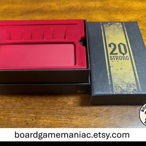 Puede incluir: Una caja de juego rectangular negra con un inserto de pl&aacute;stico rojo que presenta una ranura larga y tres compartimentos cuadrados. La tapa es negra con una franja vertical dorada y el texto 20 STRONG impreso en negro. Caja abierta.