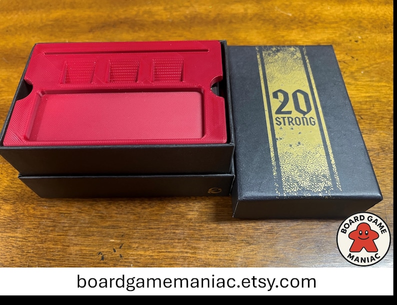 Puede incluir: Una caja de juego rectangular negra con un inserto de pl&aacute;stico rojo que presenta una ranura larga y tres compartimentos cuadrados. La tapa es negra con una franja vertical dorada y el texto 20 STRONG impreso en negro. Caja abierta.