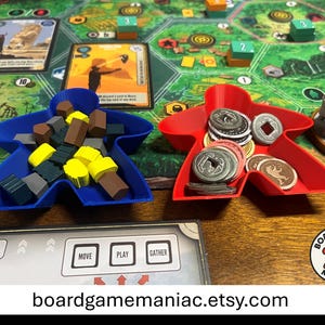 May include: Two meeple-shaped plastic trays in blue and red, used as board game component organizers. The blue tray holds various small wooden resource cubes in yellow, brown, and dark gray. The red tray contains several round metal-style game coins.