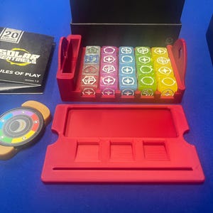 May include: A board game set with a red plastic organizer holding colorful dice and a red tray. The game includes a circular token with a multi-colored design and a rule book titled "Solar Sentinels".