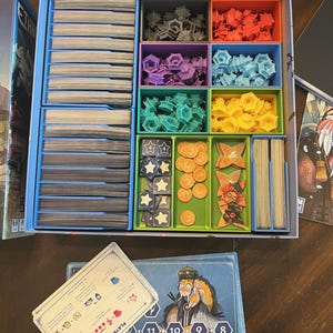 May include: A blue and white game organizer with compartments for storing game pieces and cards. The organizer has a player aid card with instructions and a player board with numbers 4 through 11.