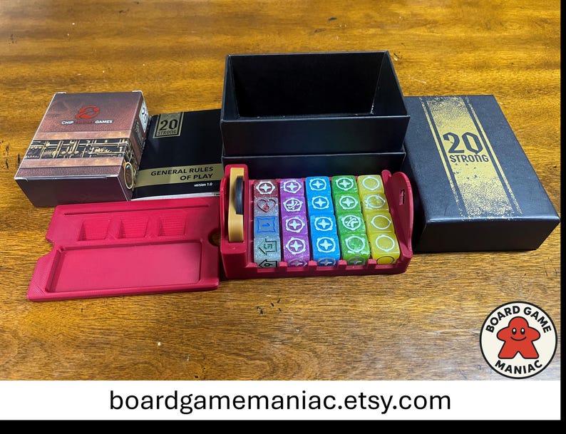 Puede incluir: Un juego de mesa 20 Strong que incluye una caja negra con texto dorado, un manual de reglas, una bandeja de pl&aacute;stico roja para dados con dados multicolores con varios s&iacute;mbolos y una bandeja de componentes roja. La tapa lleva el texto 20 STRONG.