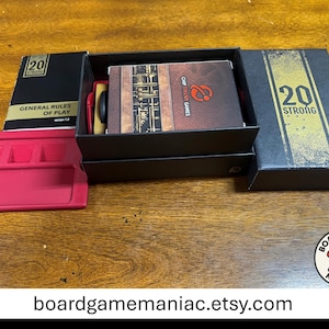 Puede incluir: Un juego de mesa 20 Strong que incluye una caja negra con texto dorado, un manual de reglas titulado General Rules of Play version 1.0, una baraja de cartas con el logo de Chip Theory Games y una bandeja de pl&aacute;stico rosa brillante con ranuras.