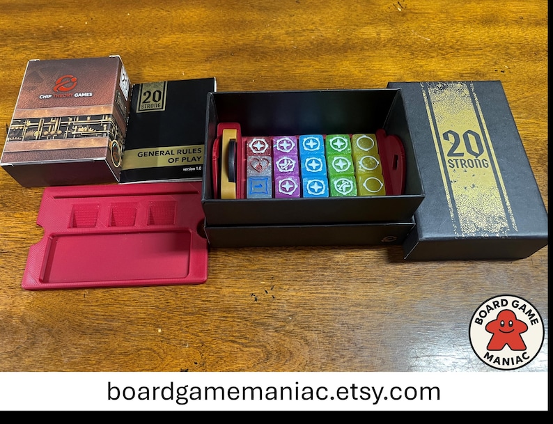 Puede incluir: Un juego de mesa 20 Strong que incluye una caja negra con texto dorado, un manual de reglas, una bandeja de pl&aacute;stico roja y una colecci&oacute;n de dados coloridos. Los dados est&aacute;n organizados en una caja de almacenamiento negra, con varios s&iacute;mbolos blancos en caras moradas, azules, verdes y amarillas. La tapa de la caja muestra el texto 20 STRONG en negrita.
