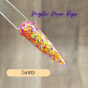 May include: A close-up of a clear nail tip with a colorful glitter polish. The glitter is a mix of orange, yellow, pink, and purple. The text "Mystic Moon Dips" is at the top of the image and "Sunrise" is at the bottom.