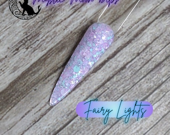 Puede incluir: Una inmersión para uñas de color morado claro y azul con purpurina sobre una base blanca. La inmersión se llama "Fairy Lights" y es de la marca "Mystic Moon Dips".