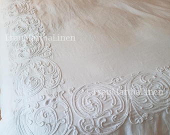 May include: A white linen duvet cover with a white embroidered design. The design features a repeating pattern of swirls and scrolls. The duvet cover has a ruffled edge.