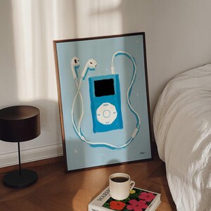 May include: A framed art print featuring a blue MP3 player with white headphones and cord. A coffee cup sits atop a book with colorful flowers and the name "WARHOL" on the cover. The artwork is displayed on a wooden floor.