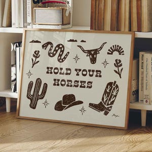 May include: A framed art print featuring a beige background and brown illustrations of a snake, longhorn skull, cactus, cowboy hat, and boot. The text "HOLD YOUR HORSES" is centered. The artwork is displayed on a white bookshelf.