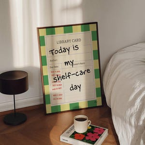Könnte beinhalten: Ein gerahmter Druck mit dem Text "Today is my shelf-care day" in einem Schlafzimmer. Eine Kaffeetasse steht auf einem Buch mit bunten Blumen. Eine kleine Lampe und ein Bett sind ebenfalls im Bild.