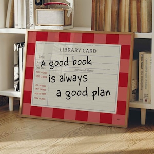 May include: A framed print with a red and white gingham border. The print resembles a library card and reads "A good book is always a good plan" in black text. The print is displayed on a wooden floor in front of a white bookshelf.