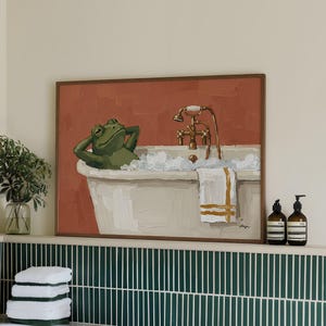 May include: A framed painting of a green frog relaxing in a white bathtub with bubbles. The artwork hangs above a white bathtub with a green towel and green tiles. Two brown bottles sit on a shelf.