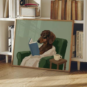 Könnte beinhalten: Ein gerahmter Kunstdruck zeigt einen braunen Dackel, der in einem grünen Sessel ein blaues Buch liest. Der Hund ist in eine weiße Decke gehüllt, mit einem kleinen Tisch und einer Tasse Kaffee daneben. Das Kunstwerk hat einen Holzrahmen.