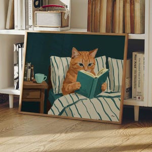 May include: A framed print showing an orange tabby cat reading a book in bed. The cat is on a bed with striped pillows and a blanket. A small side table with a blue mug is visible. The artwork is displayed in a white bookshelf setting.