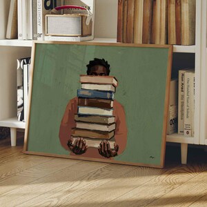 Könnte beinhalten: Ein gerahmter Kunstdruck mit einer Person, die einen Stapel Bücher hält, vor einem grünen Hintergrund. Die Person trägt ein rot-orangefarbenes Hemd. Das Kunstwerk steht auf einem Holzboden, mit einem weißen Bücherregal im Hintergrund.
