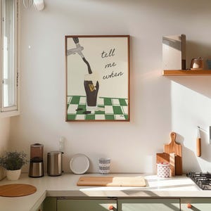 Puede incluir: Interior de cocina con un cuadro enmarcado que dice "tell me when" sobre un vaso que se llena con l&iacute;quido oscuro. La cocina tiene una combinaci&oacute;n de colores verde claro y blanco, con tablas de cortar de madera y una peque&ntilde;a planta en la encimera.