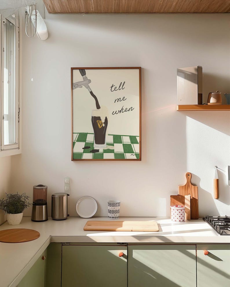 Puede incluir: Interior de cocina con un cuadro enmarcado que dice "tell me when" sobre un vaso que se llena con l&iacute;quido oscuro. La cocina tiene una combinaci&oacute;n de colores verde claro y blanco, con tablas de cortar de madera y una peque&ntilde;a planta en la encimera.