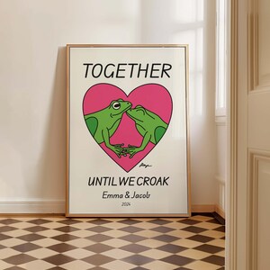 May include: A white poster with a pink heart featuring two green frogs kissing. The text "TOGETHER UNTIL WE CROAK" is below the heart, with the names "Emma & Jacob" and the year "2024" below the text.
