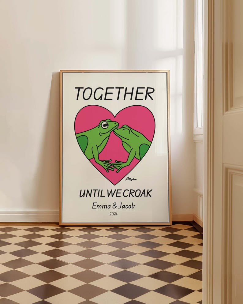 May include: A white poster with a pink heart featuring two green frogs kissing. The text "TOGETHER UNTIL WE CROAK" is below the heart, with the names "Emma & Jacob" and the year "2024" below the text.