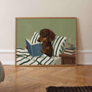 May include: A framed wall art print featuring a brown dachshund sitting up in bed with green and white striped pillows and bedding. The dog is holding an open blue book. A small wooden nightstand holds a stack of books and a steaming mug of coffee.