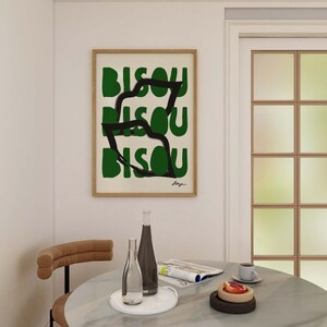 Op de afbeelding: Een ingelijst print met het woord "Bisou" dat drie keer wordt herhaald in een abstract ontwerp in groen en zwart. De print hangt aan een witte muur boven een ronde marmeren tafel met twee glazen flessen, een tijdschrift en een bord met fruit.