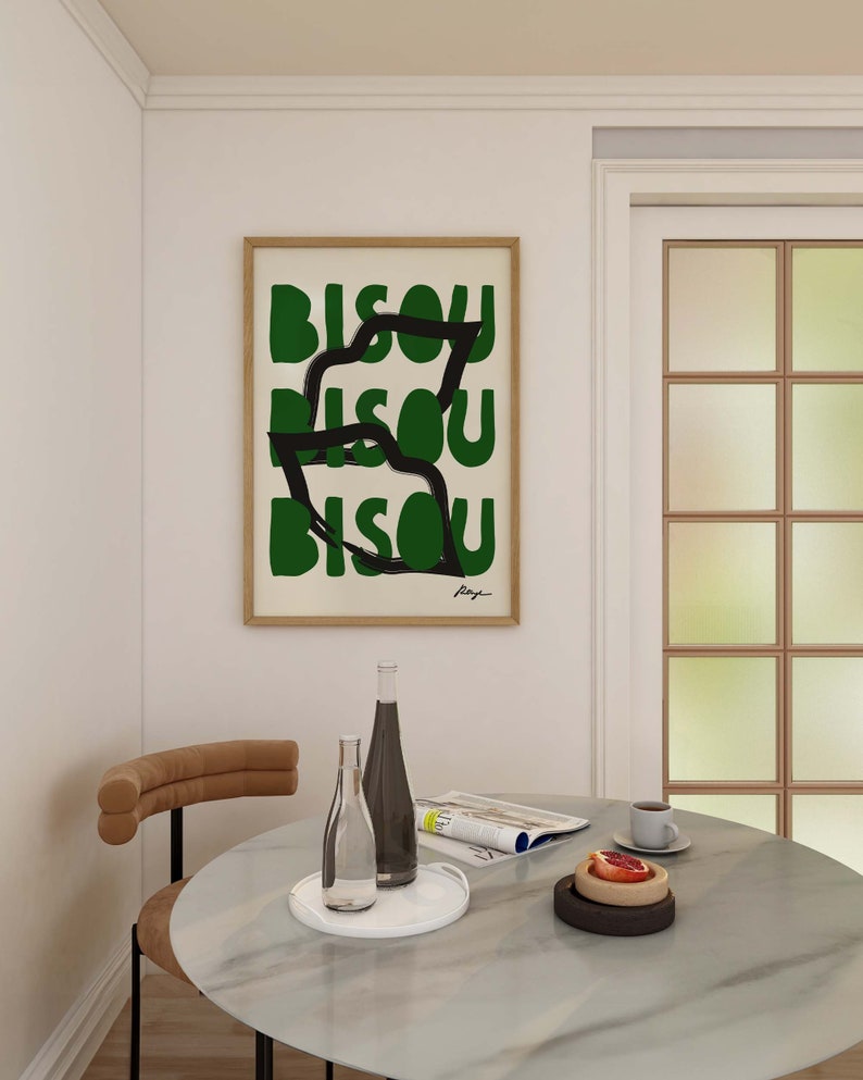 Op de afbeelding: Een ingelijst print met het woord "Bisou" dat drie keer wordt herhaald in een abstract ontwerp in groen en zwart. De print hangt aan een witte muur boven een ronde marmeren tafel met twee glazen flessen, een tijdschrift en een bord met fruit.