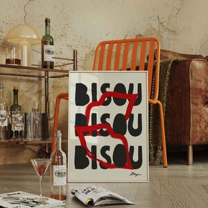 Op de afbeelding: Een ingelijst kunstprint met het woord "BISOU" dat drie keer wordt herhaald in zwarte en rode letters. De print hangt aan een muur met een houten vloer en een stoel op de voorgrond.