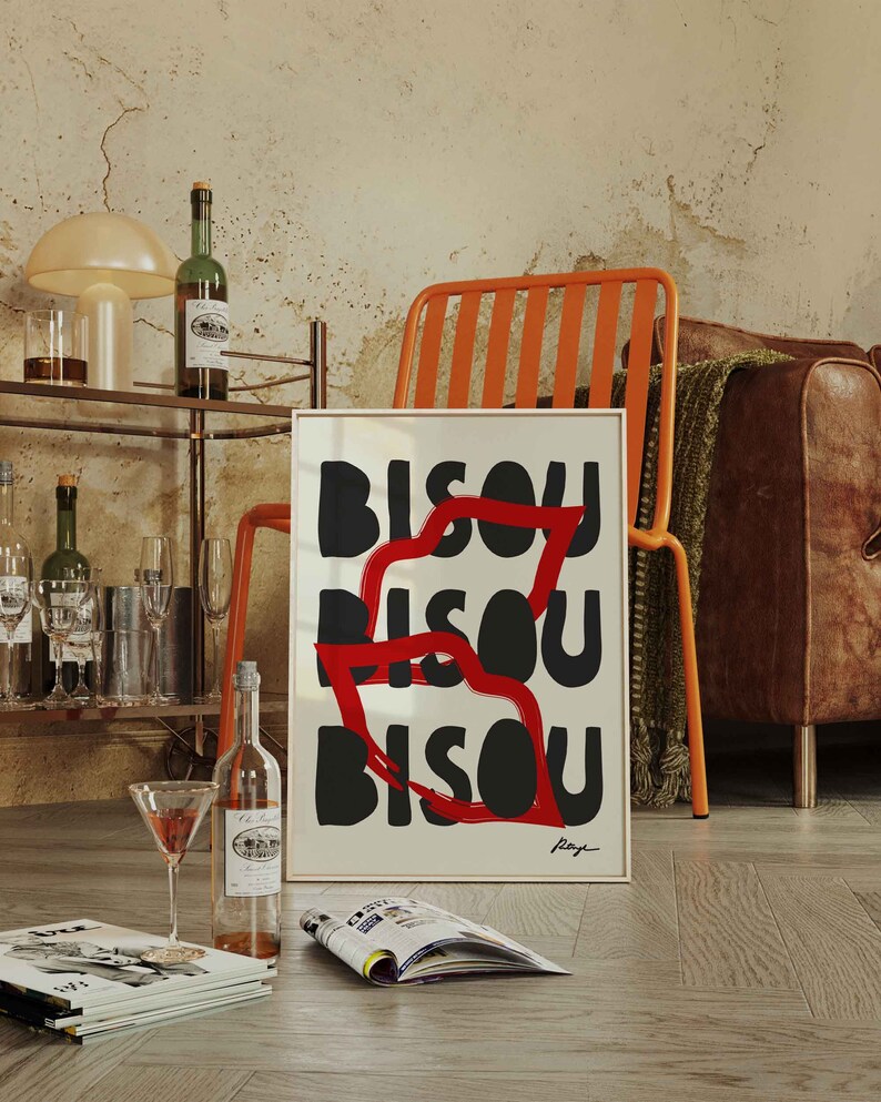 Op de afbeelding: Een ingelijst kunstprint met het woord "BISOU" dat drie keer wordt herhaald in zwarte en rode letters. De print hangt aan een muur met een houten vloer en een stoel op de voorgrond.