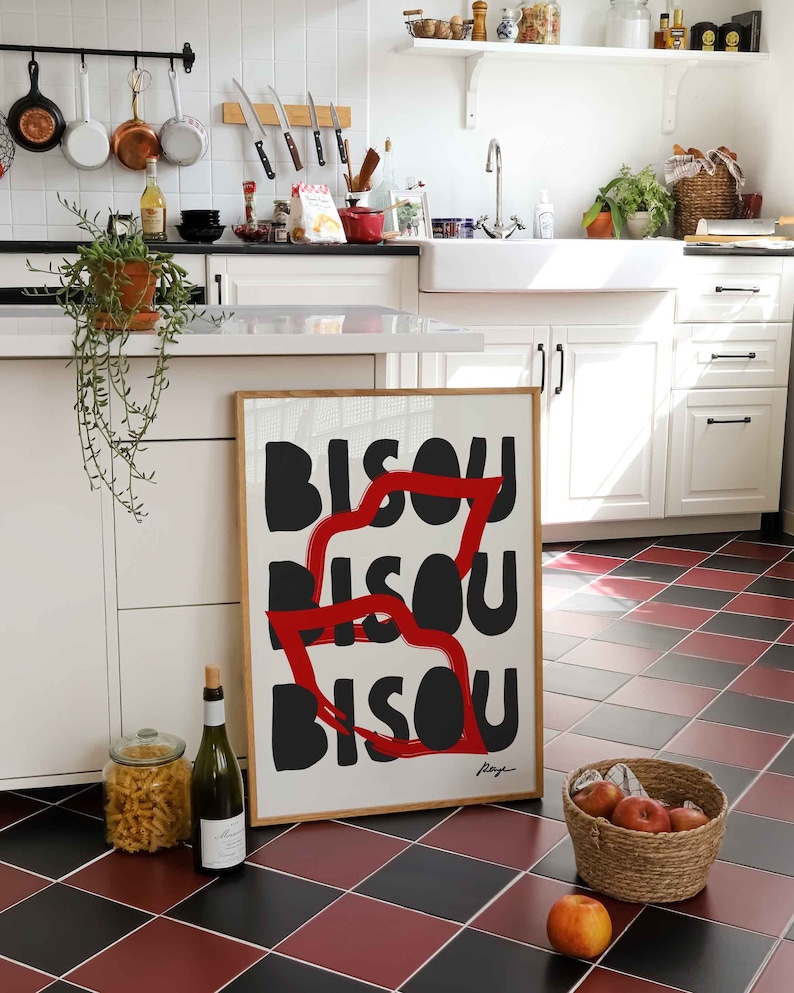 Op de afbeelding: Een ingelijst afdruk met het woord "Bisou" drie keer herhaald in zwarte tekst met een rode streep. De afdruk hangt aan een witte muur in een keuken.
