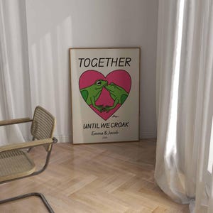 May include: A white poster with a pink heart and two green frogs kissing inside the heart. The text on the poster reads "Together Until We Croak Emma & Jacob 2024".