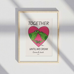 May include: A framed print with a pink heart and two green frogs kissing inside. The text reads "Together Until We Croak" with the names "Emma & Jacob" and the year "2024" below.