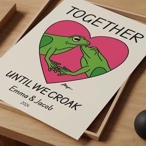 May include: A white card with a pink heart featuring two green frogs kissing. The text reads "Together until we croak - Emma & Jacob 2022".