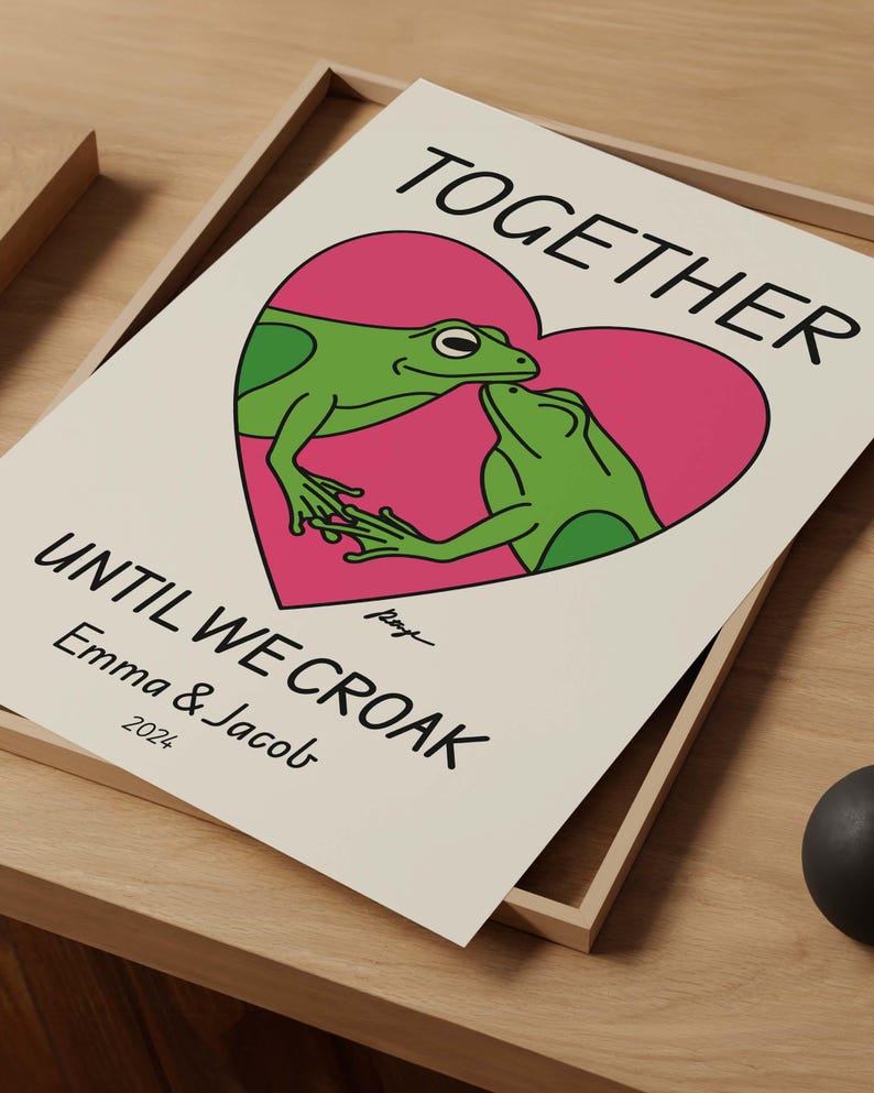 May include: A white card with a pink heart featuring two green frogs kissing. The text reads "Together until we croak - Emma & Jacob 2022".