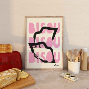 Op de afbeelding: Een ingelijst print met een roze en zwart ontwerp. De print toont het woord "Bisou" drie keer herhaald in een gestileerd lettertype. De print is rechtsonder getekend met "Raye".