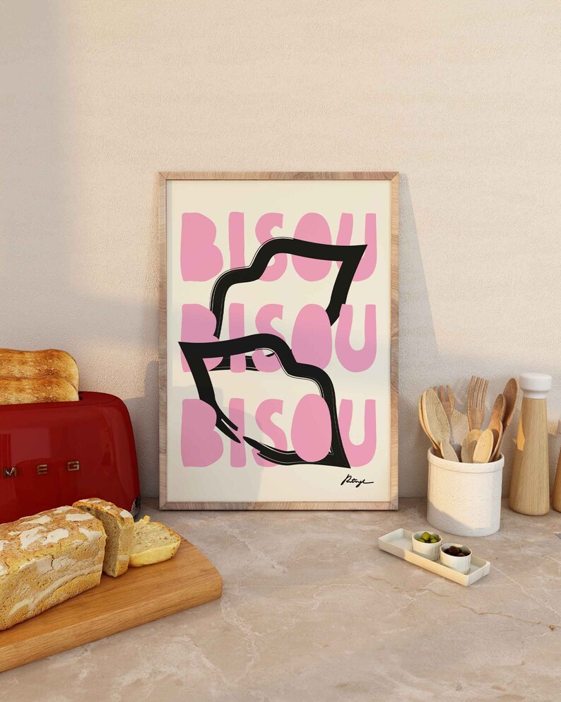 Op de afbeelding: Een ingelijst print met een roze en zwart ontwerp. De print toont het woord "Bisou" drie keer herhaald in een gestileerd lettertype. De print is rechtsonder getekend met "Raye".