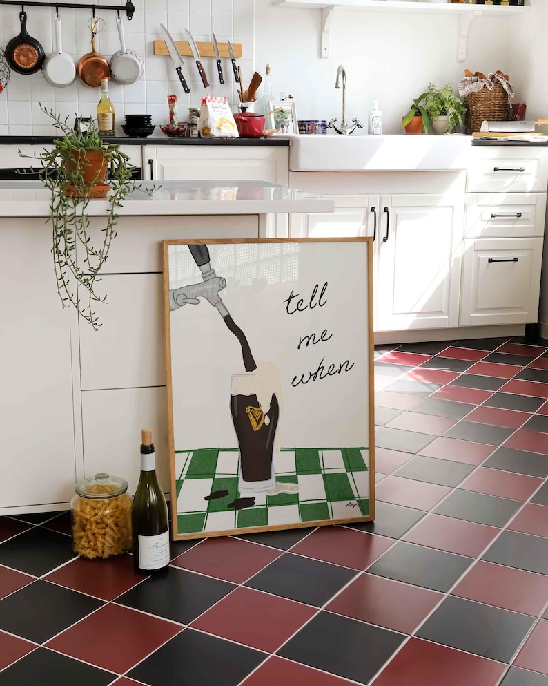 Puede incluir: Un cuadro enmarcado con las palabras "tell me when" y un dibujo de cerveza verti&eacute;ndose en un vaso. Una botella de vino y un tarro de pasta est&aacute;n frente al cuadro. El suelo tiene un patr&oacute;n de cuadros rojos y negros.