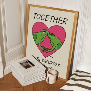 May include: A framed print with a light brown background featuring two green frogs kissing inside a pink heart. The text "TOGETHER UNTIL WE CROAK" is written below the heart.