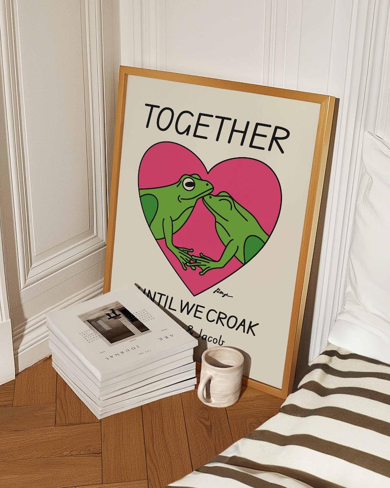 May include: A framed print with a light brown background featuring two green frogs kissing inside a pink heart. The text "TOGETHER UNTIL WE CROAK" is written below the heart.