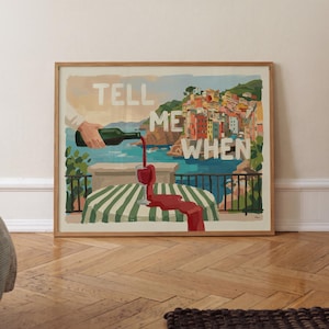 May include: A framed art print featuring a coastal scene with colorful houses, a green and white striped tablecloth, and a wine glass overflowing with red wine. The text "TELL ME WHEN" is displayed in white letters across the illustration.